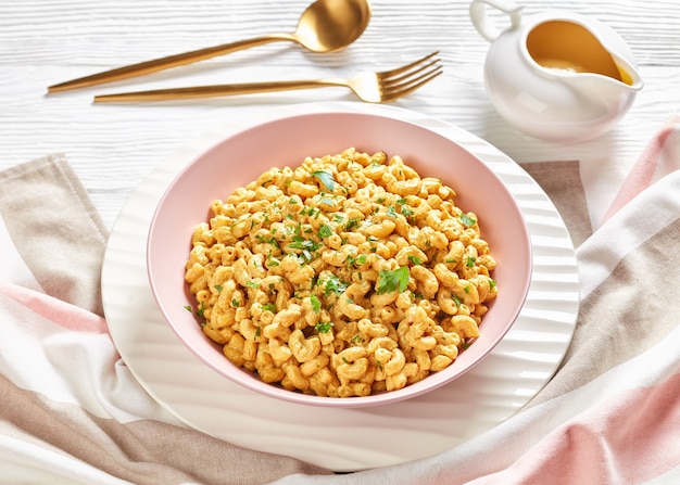 Mac et fromage végétaliens crémeux avec sauce à la levure nutritionnelle dans un bol rose sur une table en bois blanc avec couverts dorés, vue paysage d'en haut, gros plan