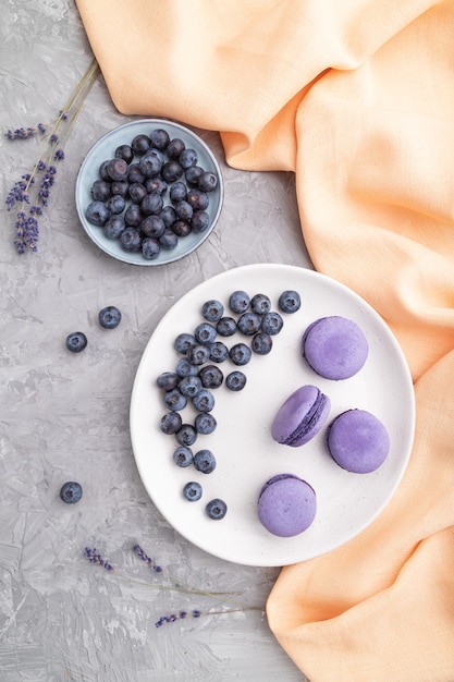 Macarons violets ou gâteaux macarons aux myrtilles sur plaque en céramique blanche sur une surface de béton gris et textile orange