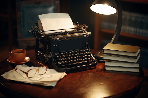 Machine à écrire vintage sur table en bois au bureau à domicile, personne. Lieu de travail de l'écrivain dans un style rétro, tasse de café et verres, lumière de la lampe