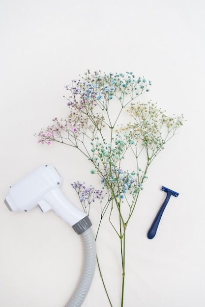 Une machine à éliminer les poils au laser et un rasoir sont juxtaposés à de délicates fleurs sur un fond clair symbolisant le contraste entre la technologie moderne et la méthode standard d'élimination des poils