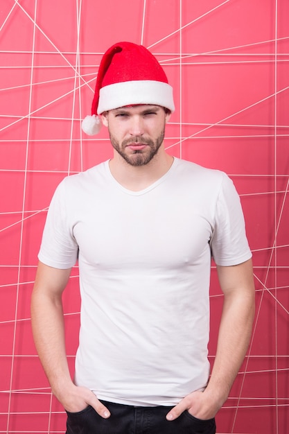 Macho en chapeau de Noël rouge et t-shirt blanc. Man santa pose sur fond rose. La mode du père noël. Fête de Noël et du nouvel an. Concept de célébration des vacances d'hiver.