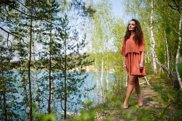 Maçonnerie en bois. Une fille aux longs cheveux bouclés ondulés vêtue d'une robe en guipure orange et de chaussures sur la nature, dans une forêt au bord du lac, se tenait près des arbres et des buissons. La jeune femme sourit et apprécie la vie