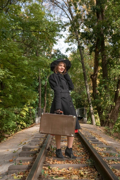 Madame se tient sur des rails avec une valise dans la forêt d'automne Femme d'âge moyen en long manteau et chapeau noir à bords