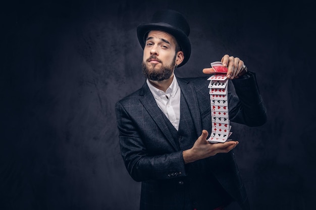 Un magicien barbu en costume noir et chapeau haut de forme, montrant un tour avec des cartes à jouer sur fond sombre.