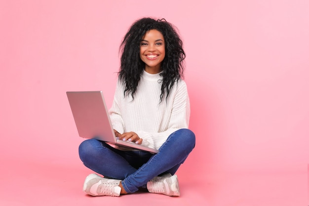 Une magnifique femme afro-américaine vêtue d'un pull blanc comme neige est assise en tailleur avec son ordinateur portable, souriant joyeusement en regardant l'écran