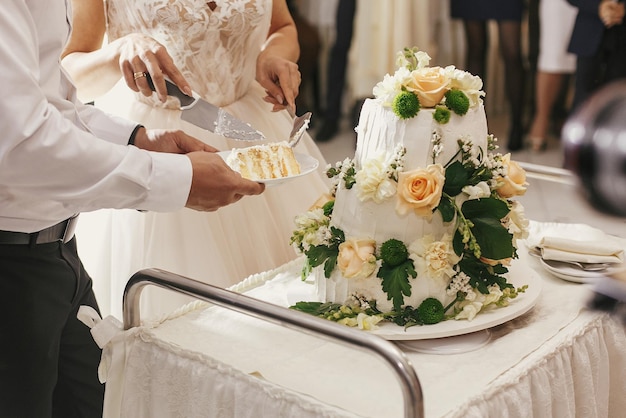 Magnifique mariée et marié élégant coupant ensemble un gâteau de mariage blanc avec des roses à la réception de mariage Gâteau de dégustation de couple de mariage heureux Moments romantiques de jeunes mariés