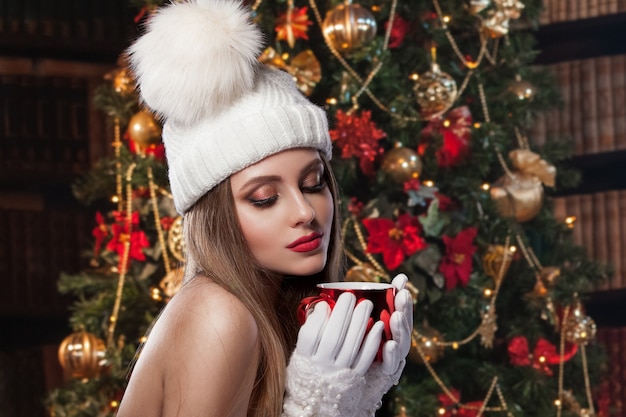 Une magnifique photo du Nouvel An. Une belle fille dans un chapeau et des mitaines tient une tasse rouge