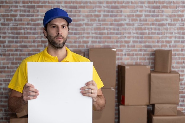 Mailman brésilien tenant un tableau vierge dans un dépôt avec beaucoup de boîtes. Copiez l'espace.