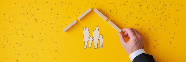 Main d'un agent d'assurance abritant une silhouette en papier découpé d'une famille en construisant un toit de chevilles en bois dans une image conceptuelle de l'assurance et de l'immobilier.