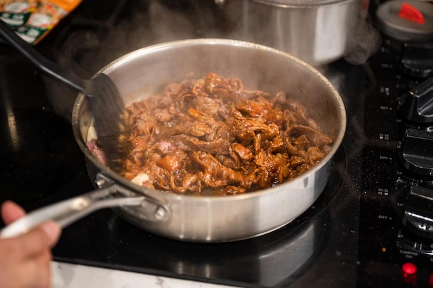 Main à l'aide de flipper de cuisine faisant frire du boeuf en tranches dans une poêle en fer sur une cuisinière électrique