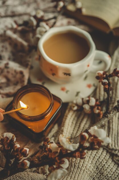 La main allume une bougie à côté d'une tasse de thé dans une tasse florale avec des fleurs de printemps et des textures confortables dans une atmosphère chaleureuse et relaxante