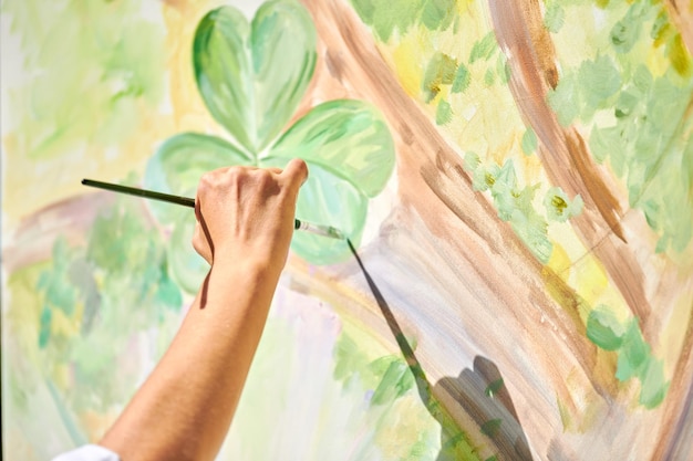 La main de l'artiste fille tient un pinceau et dessine un paysage de nature verdoyante sur toile