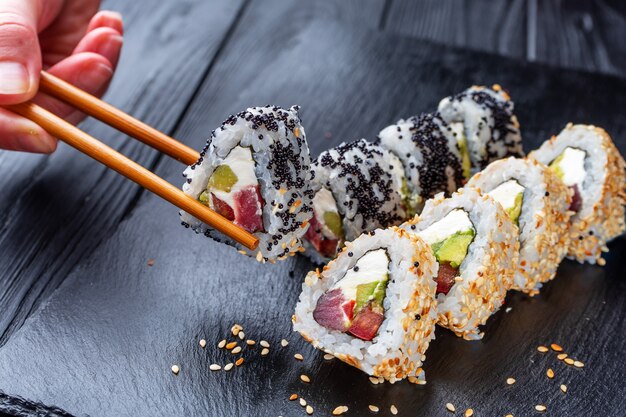 La main avec des baguettes prend des sushis à partir de rouleaux de sushi avec du fromage à la crème, du riz et du saumon sur un tableau noir décoré de gingembre et de wassabi sur une table en bois sombre. Nourriture japonaise. Flou sélectif