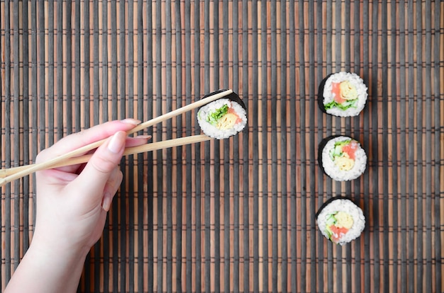 Une Main Avec Des Baguettes Tient Un Rouleau De Sushi Sur Un Mur De Tapis En Paille De Bambou. Cuisine Asiatique Traditionnelle.