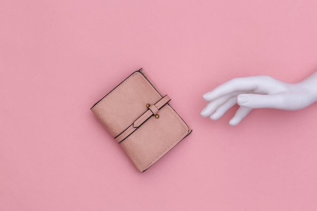 La main blanche du mannequin touche le portefeuille en cuir sur fond rose. Vue de dessus
