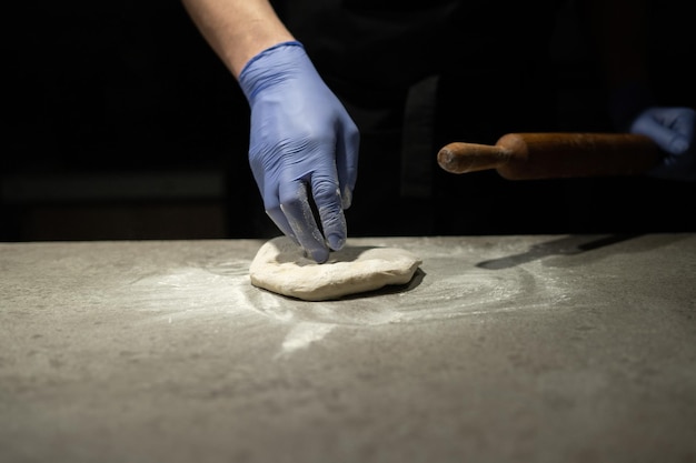 La main d'un boulanger dans un gant en caoutchouc pétrit la pâte à pizza sur la table de travail. Rouleau à pâtisserie en bois à la main. Processus de cuisson.