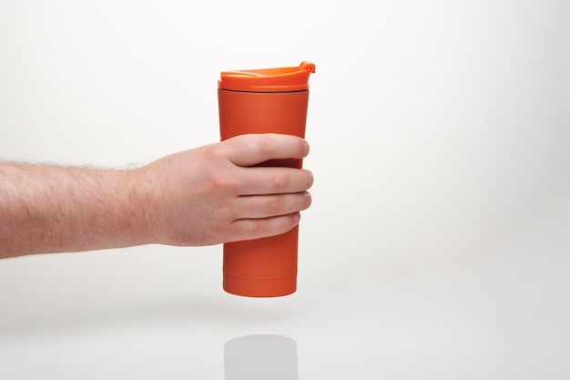 La main brutale de l'homme tient la tasse de voyage orange. Tasse à café réutilisable à emporter. Bouteille thermos en acier inoxydable avec couvercle à glissière. Maquette de tasse pour boissons froides et chaudes isolées sur fond blanc.