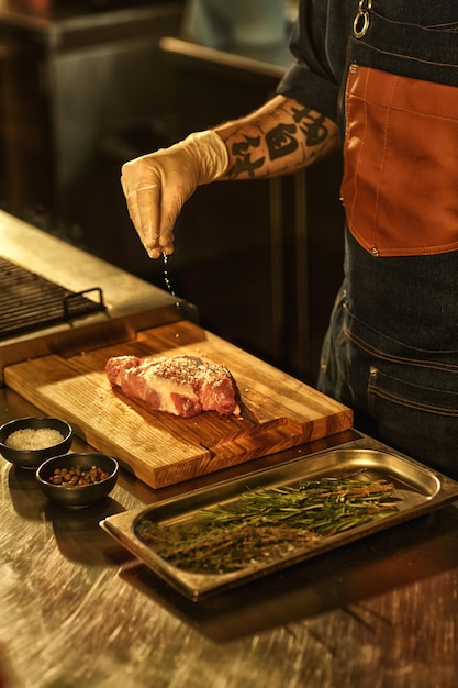 Photo main de chef professionnel en tablier et gant blanc salant le steak de boeuf à la viande crue sur une planche à découper en bois avec assaisonnements et romarin concept de cuisine de restaurant culinaire et cuisine