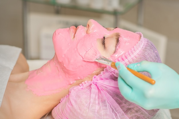 Main de cosmétologue appliquant un masque alginique rose au visage de la jeune femme