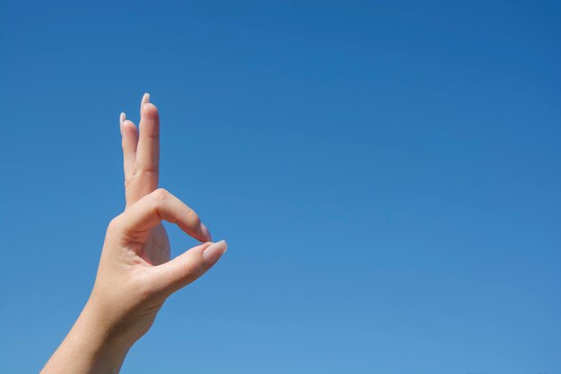 La main coupée d'une femme faisant un signe ok contre un ciel bleu clair.