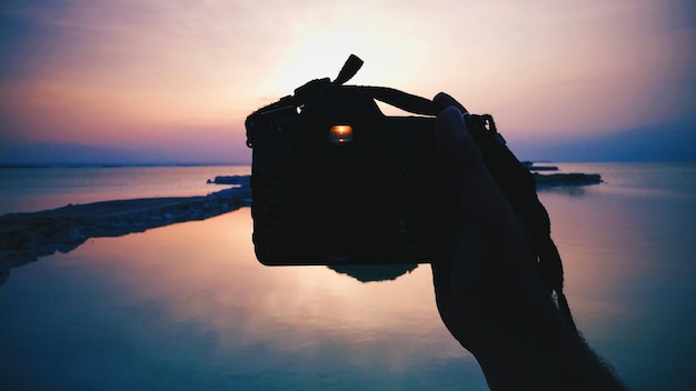 Photo main coupée d'un homme photographiant la mer contre un ciel nuageux au coucher du soleil à partir d'un appareil photo