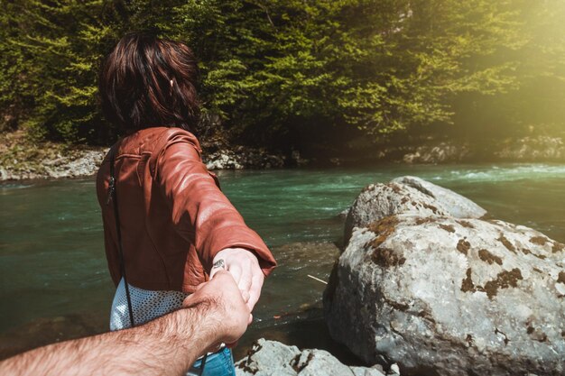 Photo main coupée d'un homme tenant la main de sa petite amie au bord d'une rivière dans la forêt