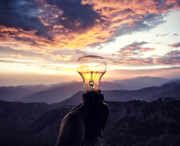 Photo main coupée d'une personne tenant une ampoule contre les montagnes et le ciel au coucher du soleil