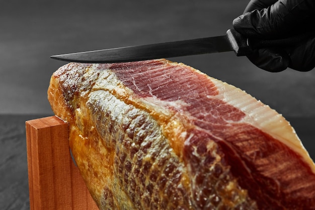 La main d'un cuisinier dans un gant noir découpe un morceau de jambon avec un couteau d'une cuisse de porc sur un stand de jamonera Gros plan Vue latérale