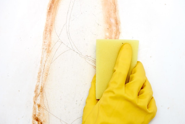 La main dans un gant jaune avec une éponge lave la rouille avec un bain émaillé blanc en gros plan