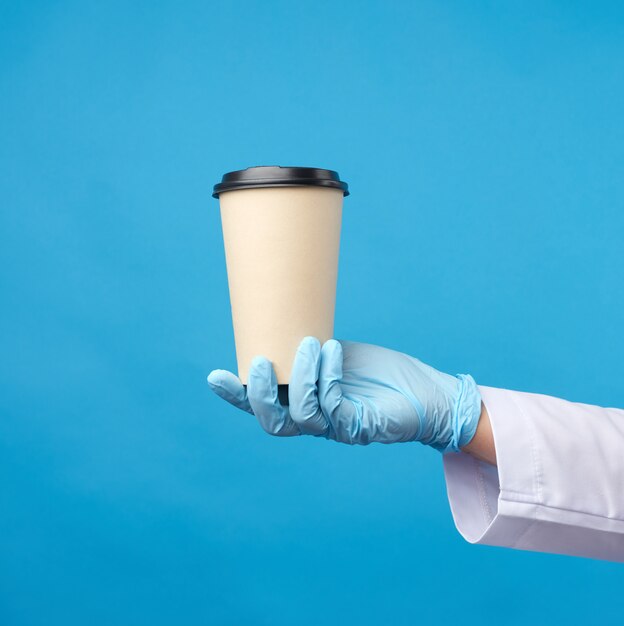 La main dans un gant en latex bleu tient une tasse en carton jetable en papier et une couverture en plastique