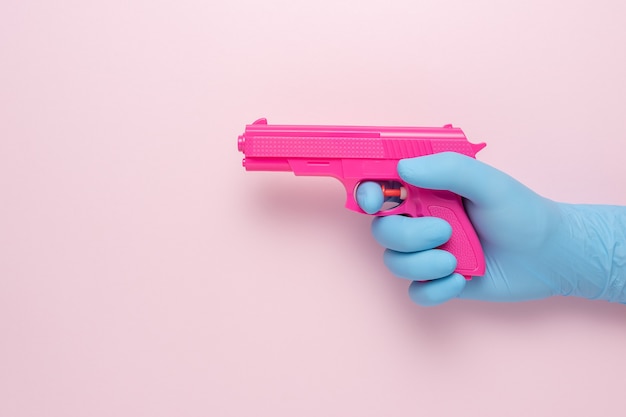 Main dans un gant médical avec une arme de poing rose sur fond rose