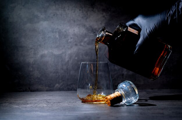 Photo la main dans un gant noir verse le whisky d'une bouteille carrée dans un verre sur une surface de ciment gris foncé