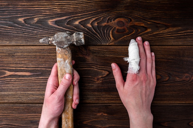 La main avec un doigt blessé tient un marteau sur une table en bois sombre.