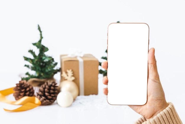 La main droite de la femme tient un smartphone avec un écran blanc isolé sur le fond d'arbres de Noël boîtes-cadeaux boules de Noël et ruban doré Il y a un chemin de découpage Concept de jour important