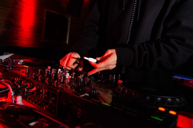 La main du DJ masculin tourne les gobelets et tient la cigarette électronique. Photo en gros plan. Notion de vie nocturne. Homme jouant de la musique à la fête en boîte de nuit. Ambiance sombre.