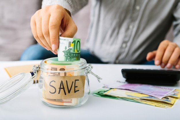 La main du peuple mettant des billets en euros dans une bouteille en verre pour économiser. L'homme fait sa comptabilité.