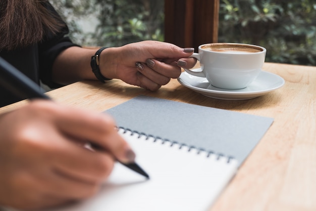 Une main écrit sur un cahier vierge blanc tout en buvant du café sur une table en bois