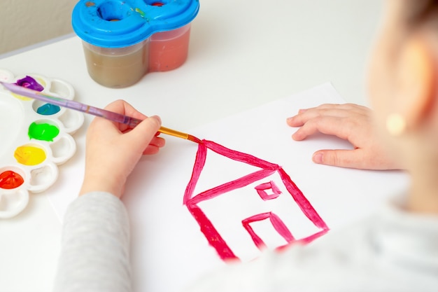 La main de l'enfant avec un pinceau dessinant la maison sur du papier blanc à l'aquarelle.