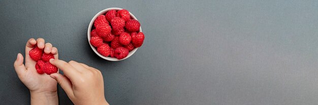 La main de l'enfant prend des framboises fraîches et juteuses dans une petite assiette noire. Gros plan rouge vif pourpre. Temps de cueillette des baies d'été. Fruits biologiques sains pour les enfants.