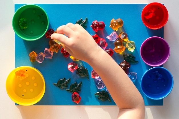 Photo main d'enfant séparant par couleur des pierres jouets multicolores éparpillées sur la surface bleue dans des tasses colorées