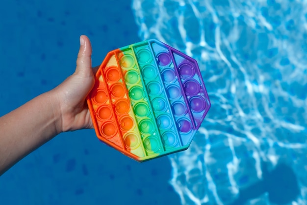 La main de l'enfant tient le jouet pop-it anti-stress coloré en silicone populaire au-dessus de l'eau dans la piscine