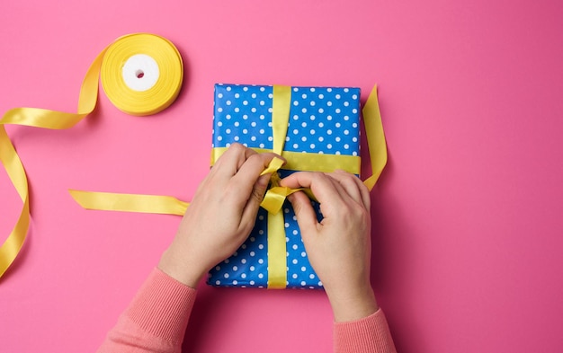 La main féminine attache un ruban de soie sur une boîte-cadeau, fond rose. Préparation des vacances