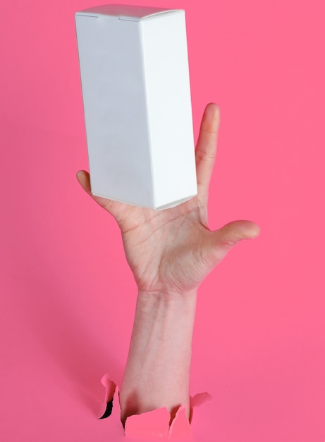 Une main féminine attrape une boîte blanche à travers du papier rose déchiré. Concept créatif minimaliste
