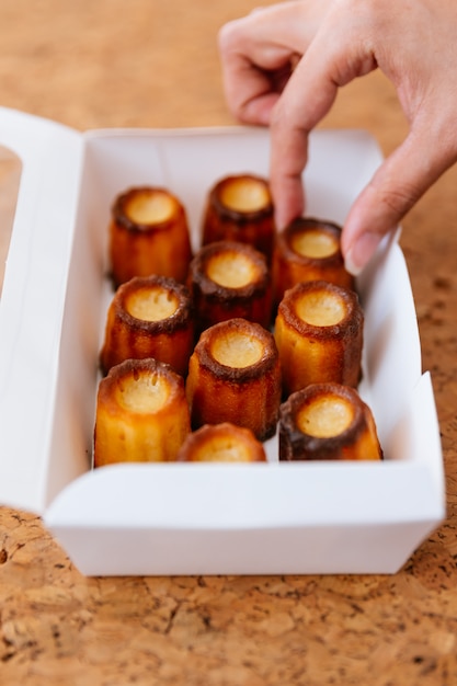 Une main féminine cueille des canelés fraîchement cuits dans une boîte en papier blanc.