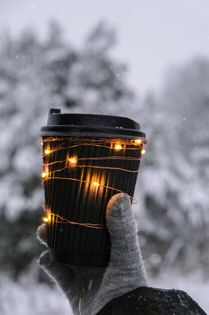 Main féminine dans des gants chauds d'hiver tenant une tasse en papier écologique noir avec une lumière brillante de guirlande Tasse de recyclage zéro déchet à la mode créative Maquette Boisson chaude chaude à emporter en hiver Concept à emporter