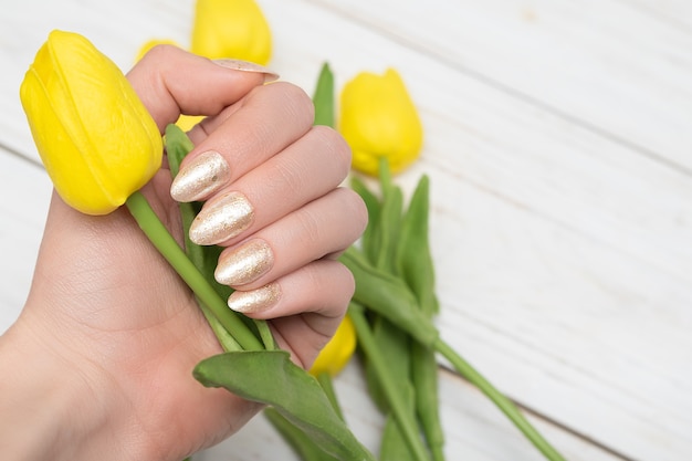 Main féminine avec un design d'ongle doré. Manucure vernis à ongles or pailleté. Main de femme tenir des tulipes de printemps jaunes sur fond de bois blanc. Espace de copie