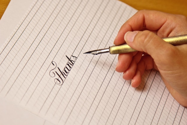 Main féminine écrit avec le stylo à encre le mot merci sur une feuille de papier blanc avec des rayures papeterie sur le bureau gros plan vue de dessus leçons d'orthographe et exercices de caligraphie arrière-plan de mise en page du modèle