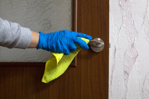 Main féminine en gant de caoutchouc bleu essuie la poignée de porte avec une serviette jaune. Désinfection de la poignée de porte de la maison