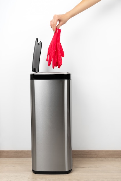 Une main féminine jette un gant de protection rose dans la poubelle sur un fond gris.