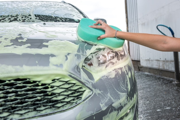 Main féminine avec de la mousse de savon éponge lave sa voiture à un lave-auto en libre-service à l'extérieur